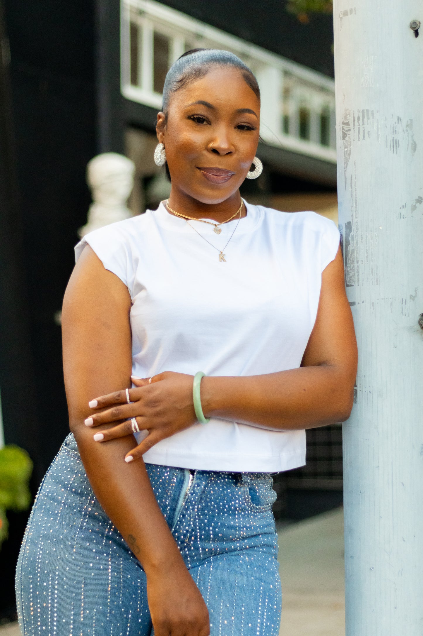 A woman leans against a light-colored pole outdoors, wearing a white short-sleeve crop top and blue jeans with small white speckles. She is accessorized with a green bracelet, hoop earrings, and a necklace.