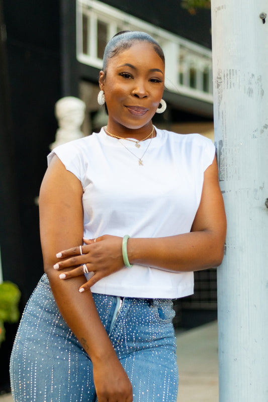A woman leans against a light-colored pole outdoors, wearing a white short-sleeve crop top and blue jeans with small white speckles. She is accessorized with a green bracelet, hoop earrings, and a necklace.